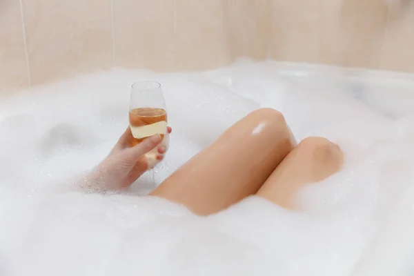 Foto da vicino di giovane donna sexy con corpo nudo dritto e gambe pulite sdraiate e rilassanti nella vasca da bagno in schiuma bianca con candele in giro in bagno leggero, bere alcol dal bicchiere di vino all'interno . — Foto Stock