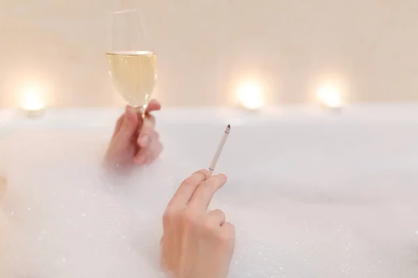 Foto da vicino di giovane donna sexy con la pelle pulita salute che giace nella vasca da bagno in schiuma bianca con candele in giro in bagno leggero, bere alcol dal bicchiere di vino e fumare sigarette all'interno . — Foto Stock