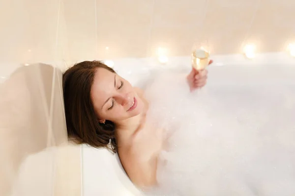 Schöne junge sexy Frau mit langen braunen Haaren und glattem nackten Körper liegend und entspannend in weißer Schaumbadewanne mit Kerzen herum im hellen Badezimmer, trinken Alkohol aus Weinglas drinnen. — Stockfoto