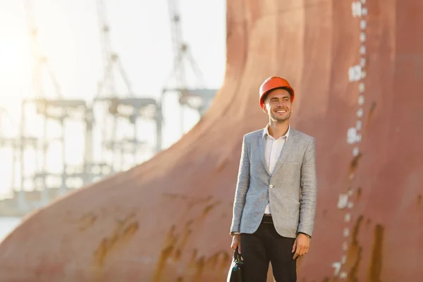 Handsome young unshaven successful business man in gray suit and protective construction orange helmet holding case, standing in sea port against a cargo rusty ship with water line background