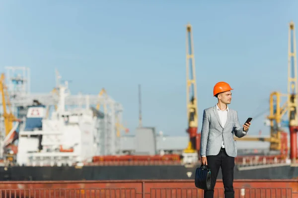 Handsome young unshaven successful business man in gray suit and protective construction orange helmet holding case, looking on mobile phone, walking in sea port against cargo ship, crane background.