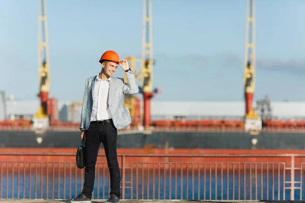 Handsome young unshaven successful business man in gray suit and protective construction orange helmet holding case, walking in sea port against cargo ship and crane background in summer time.