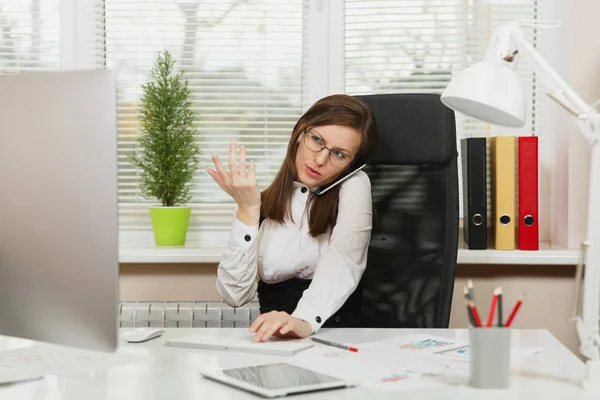 Beautiful serious business woman in suit and glasses sitting at the desk with tablet, working at computer with documents in light office, talking on mobile phone resolving issues, looking aside — Stock Photo, Image