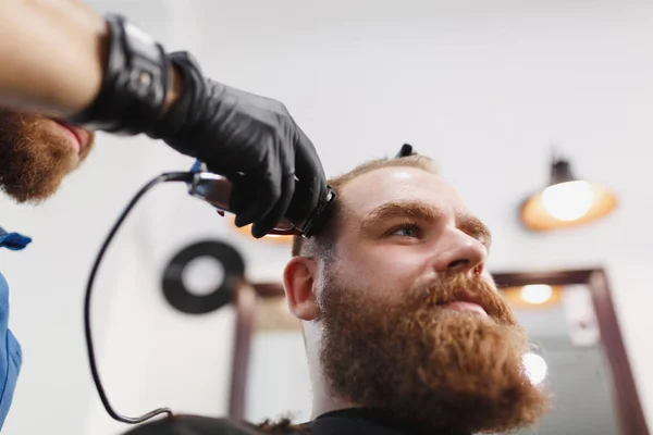 Close up of male professional hairdresser serving client by clipper. Ginger handsome stylish young man with big beard and short hair getting trendy haircut in black cape. Light white barber shop room. — Stock Photo, Image