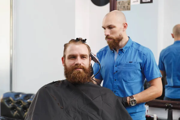 Мужчина профессиональный парикмахер, обслуживающий клиента клиппером. Рыжий красивый грубый стильный молодой человек с густой бородой и короткими волосами, стригущийся в черном плаще. Светло-белая парикмахерская — стоковое фото