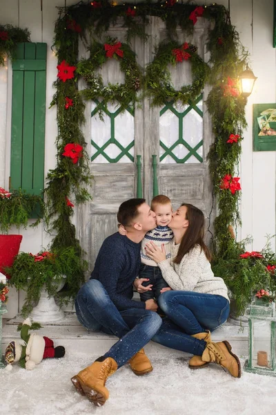 Happy parents kissing little son. Child boy in sweater sitting on porch snow steps at light house with decorated in red green New Year door at home. Christmas good mood. Family, holiday 2018 concept — Stock Photo, Image