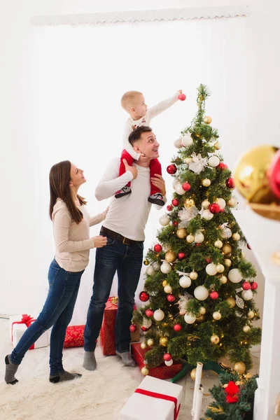 Feliz joven padres alegres con lindo hijo pequeño. Niño niño sobre los hombros papi decorando árbol de Año Nuevo con bola de juguete en el cuarto de luz en casa. Navidad buen humor. Familia, amor y vacaciones 2018 concepto — Foto de Stock