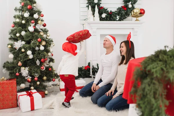 Happy young cheerful parents fighting pillows with cute little son. Child boy in red hat in light room at home with decorated New Year tree. Christmas good mood. Family, love and holiday 2018 concept. — Stock Photo, Image