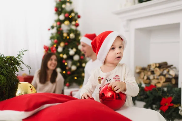 Padres jóvenes felices con lindo hijo en sombrero. Niño con gran bola de juguete de árbol rojo de pie en la sala de luz decorada de Año Nuevo en casa. Navidad buen humor. Familia, amor y vacaciones 2018 concepto . — Foto de Stock