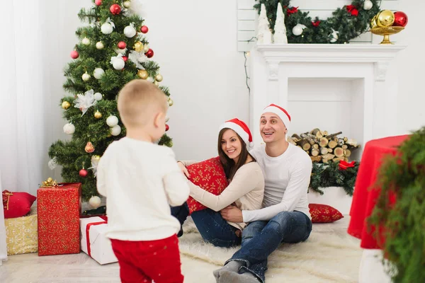 Jovens pais alegres felizes com o filhinho bonito em chapéu vermelho. Menino na sala de luz em casa com árvore de Ano Novo decorada e caixas de presente. Bom humor de Natal. Conceito de família, amor e férias 2018 . — Fotografia de Stock