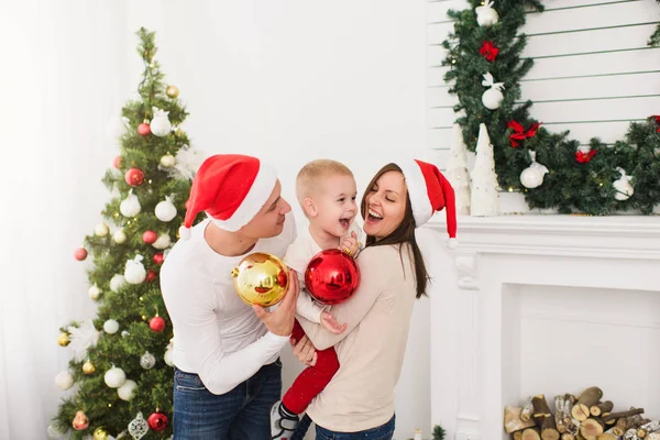 Gelukkig vrolijke ouders met schattige zoontje. Kind jongen met grote rode boom speelgoed bal staande in ingerichte Nieuwjaar lichte kamer thuis op de achtergrond van de kroon van Kerstmis. Familie, concept vakantie 2018. — Stockfoto