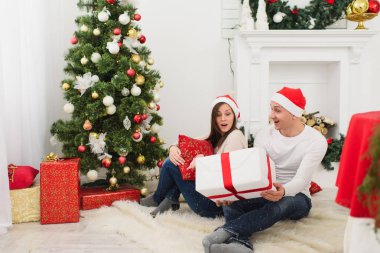 Mutlu neşeli komik genç çift dekore edilmiş yeni yıl ağacı ve hediye kutuları arka planda Noel çelenk ışık Oda evde oturan kırmızı şapkalı aşık. Aile, tatil 2018 kavramı.