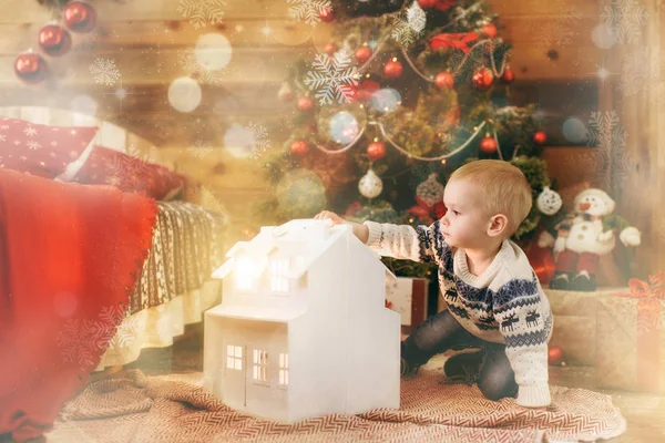 Niño feliz sentado en el árbol de Navidad con una casa blanca de juguete en la habitación de madera en casa. Buen humor para niños. Año Nuevo. Estilo de vida, familia y vacaciones 2018 concepto. Resalte mágico efecto bokeh — Foto de Stock