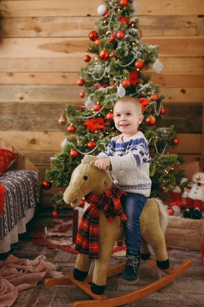 Szczęśliwy ładny mały chłopczyk na koń na biegunach na choinkę z zabawką, ubrany w sweter i dżinsy w urządzonych pokojach nowy rok w domu. Dziecko bardzo dobry nastrój. Styl życia, rodziny i wakacje koncepcja 2018 — Zdjęcie stockowe