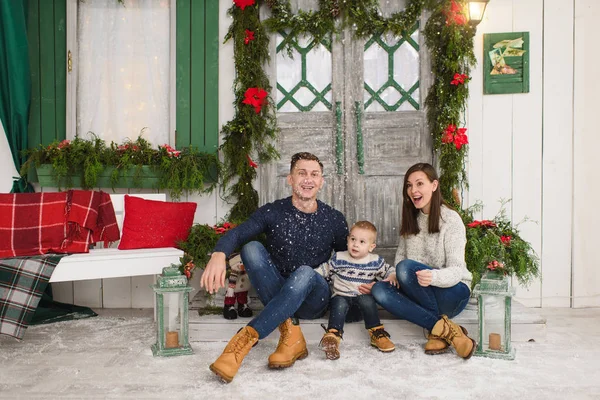 Happy parents with little son. Child boy in sweater sitting on porch snow steps at light house with decorated in red green New Year door at home. Christmas good mood. Family and holiday 2018 concept — Stock Photo, Image