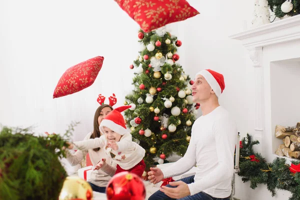 Happy young cheerful parents fighting pillows with cute little son. Child boy in red hat in light room at home with decorated New Year tree. Christmas good mood. Family, love and holiday 2018 concept. — Stock Photo, Image