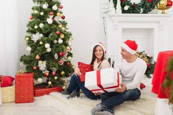Happy cheerful funny young couple in love in red hat sitting in light room at home with decorated New Year tree and gift boxes on the background of Christmas wreath. Family, holiday 2018 concept.