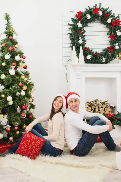 Feliz alegre casal jovem engraçado no amor em chapéu vermelho sentado na sala de luz em casa com árvore de Ano Novo decorado e caixas de presente no fundo da grinalda de Natal. Família, férias 2018 conceito . — Fotografia de Stock