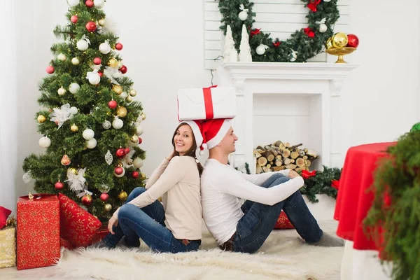 Gelukkig vrolijke grappige jonge verliefde paar in rode hoed zitten in lichte kamer thuis met ingerichte nieuwe jaar boom cadeau vakken en op de achtergrond van de kroon van Kerstmis. Familie, concept vakantie 2018. — Stockfoto