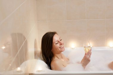 Uzun kahverengi saçlı ve düz çıplak vücut yalan, beyaz köpük küvet mum ışık banyoda, şarap cam kapalı içki alkol ile rahatlatıcı olan güzel seksi kadın.