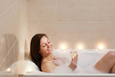 Uzun kahverengi saçlı ve düz çıplak vücut yalan, beyaz köpük küvet mum ışık banyoda, şarap cam kapalı içki alkol ile rahatlatıcı olan güzel seksi kadın.