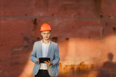 Yakışıklı tıraşsız başarılı iş adamı gri takım elbise, tablet, liman kargo paslı gemi arka plan ayakta tutan koruyucu inşaat turuncu kask. Erkek ile güneşli günde becerikli alet