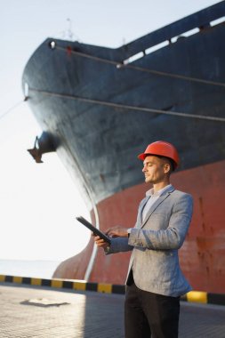 Yakışıklı tıraşsız başarılı iş adamı gri takım elbise, tablet, liman kargo paslı gemi arka plan ayakta tutan koruyucu inşaat turuncu kask. Erkek ile güneşli günde becerikli alet