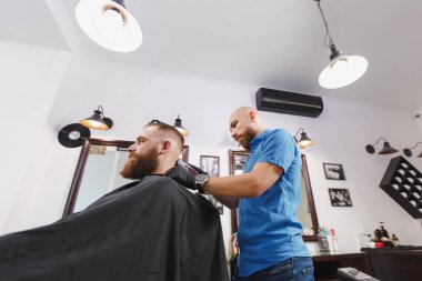 Erkek profesyonel kuaför hizmet istemci tarafından kesme makinesi. Zencefil acımasız şık yakışıklı genç kalın büyük sakal ve kısa saç Siyah pelerinli trendy saç kesimi almak. Işık beyaz Kuaför Salonu Oda