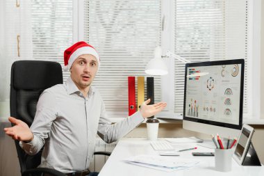 Çalışma ile modern sınıf başkanı bilgisayar, pencere arka plan üzerinde ışık office belgelerinde, eller, yayılan yakışıklı şok ve çapraşık iş adamı masa başında oturan gömlek ve kırmızı Noel şapkalı