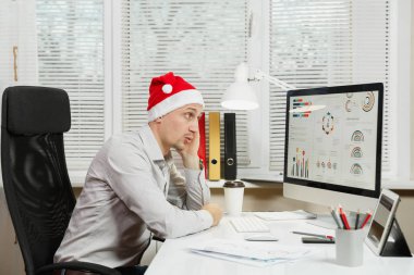 Pencere arka plan üzerinde ışık Office belgeleri ile modern monitör bilgisayarda çalışan yakışıklı şok ve çapraşık iş adamı masa başında oturan gömlek ve kırmızı Noel şapkalı. Yeni yıl.
