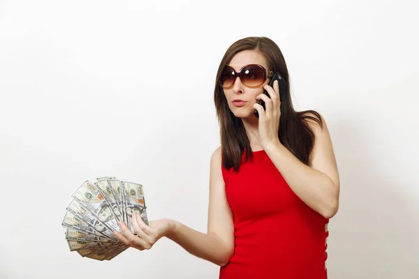 Jolie jeune femme d'affaires caucasienne heureuse en robe rouge et lunettes parlant sur téléphone portable, tenant des billets d'argent sur fond blanc. Belle fille avec argent regardant de côté isolé . — Photo