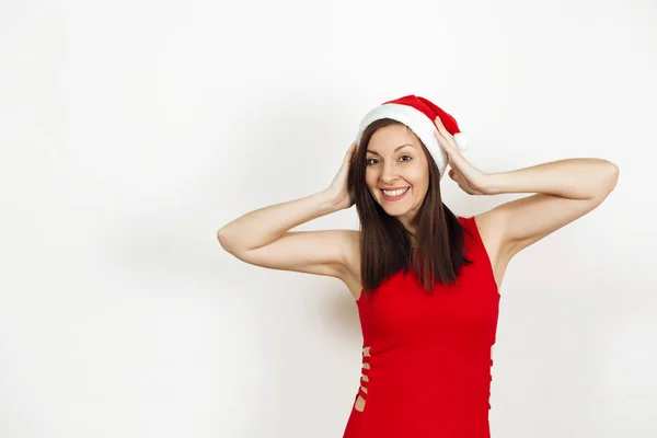 Ganska kaukasiska ung glad kvinna med frisk hud och charmiga leende bär röd klänning och jul hatt håller händerna på huvudet isolerad på vit bakgrund. Santa flicka. Nytt år holiday 2018 — Stockfoto