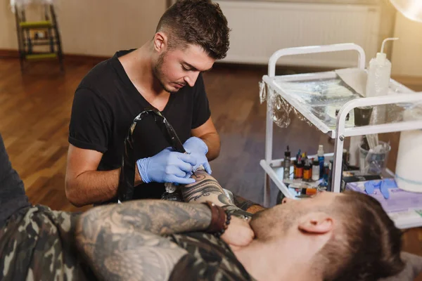 Professional tattooer artist doing picture on hand of man by machine black ink from a jar. Tattoo art on body. Equipment for making tattoo art. Master makes tattooed in light studio.