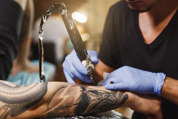 Close up of professional tattooer artist doing picture on hand of man by machine black ink from a jar. Tattoo art on body. Equipment for making tattoo art. Master makes tattooed in light studio.