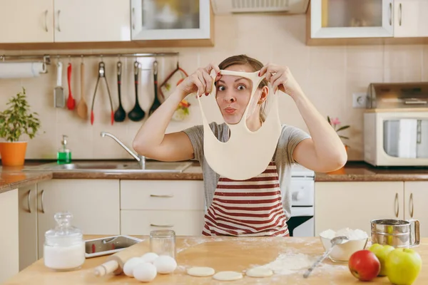 Eine junge lustige, fröhliche und lächelnde Frau legt einen Teig mit Löchern im Gesicht an und hat Spaß in der Küche. Kochen zu Hause. Essen zubereiten. — Stockfoto