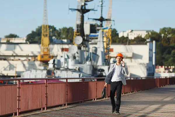 Handsome young unshaven successful business man in gray suit and protective construction orange helmet holding case, talking on mobile phone, walking in sea port against cargo ship, crane background