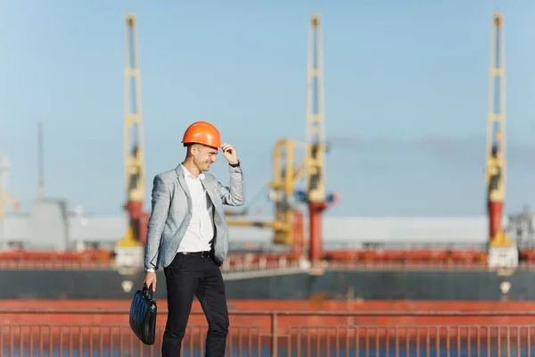 Handsome young unshaven successful business man in gray suit and protective construction orange helmet holding case, walking in sea port against cargo ship and crane background in summer time.