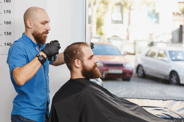 Male professional hairdresser serving client by clipper. Ginger handsome brutal stylish young man with thick big beard and short hair getting trendy haircut in black cape. Light white barber shop room