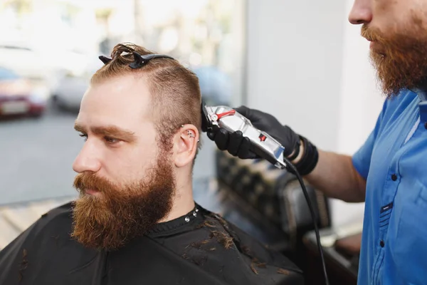 Закрытие профессионального парикмахера-мужчины, обслуживающего клиента клиппером. Рыжий красивый стильный молодой человек с большой бородой и короткими волосами, стригущийся в черном плаще. Светло-белая парикмахерская . — стоковое фото