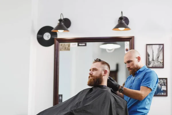 Männliche professionelle Friseurin, die Kunden per Clipper bedient. Ingwer gutaussehender brutaler, stylischer junger Mann mit dickem großen Bart und kurzen Haaren, der sich eine trendige Frisur in schwarzem Umhang schneidet. heller weißer Friseursalon — Stockfoto