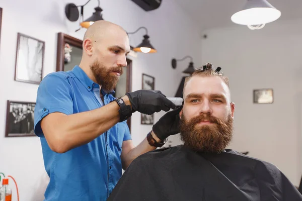 Męski profesjonalny salon fryzjerski klienta przez clipper. Ginger przystojny brutalny stylowe młody człowiek z brody duże grube i krótkie włosy coraz modne fryzury w czarnej pelerynie. Światło białe barber shop Pokój — Zdjęcie stockowe