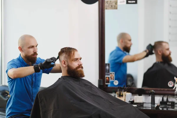 Męski profesjonalny salon fryzjerski klienta nożycami. Ginger przystojny brutalny stylowe młody człowiek z brodą duże grube, krótkie włosy coraz modne fryzury w czarnej pelerynie. Światło białe barber shop Pokój. — Zdjęcie stockowe