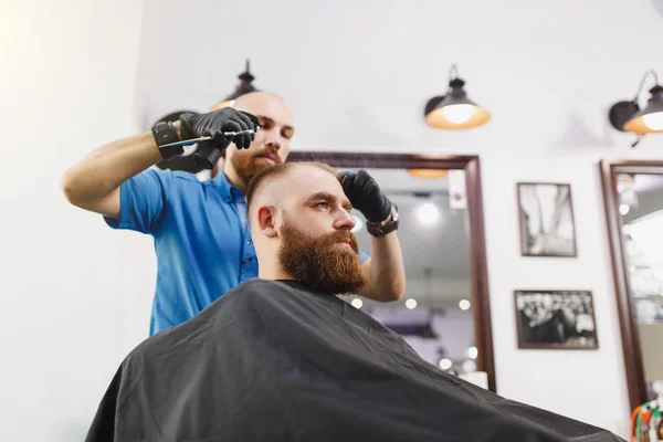 Cabeleireiro profissional masculino servindo cliente por tesoura. Gengibre bonito jovem elegante brutal com barba grande grossa, cabelo curto ficando moderno corte de cabelo em capa preta. Sala de barbeiro branco claro . — Fotografia de Stock