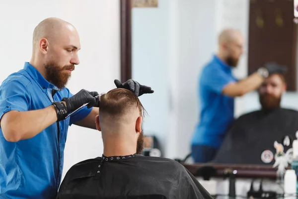 Male professional hairdresser serving client by scissors. Ginger handsome brutal stylish young man with thick big beard, short hair getting trendy haircut in black cape. Light white barber shop room.