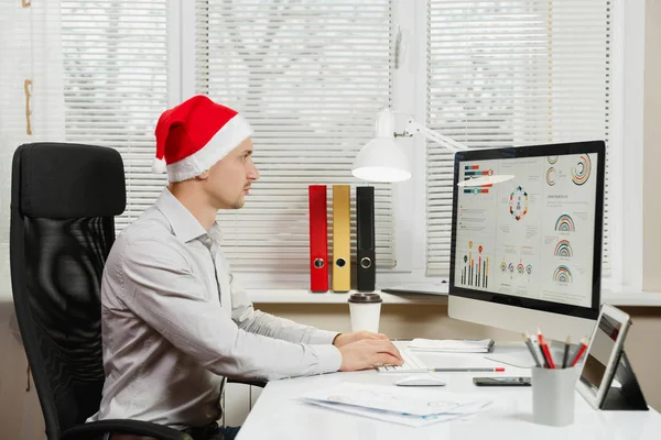 Hombre de negocios guapo serio y absorto en camisa y sombrero rojo de Navidad sentado en el escritorio, trabajando en la computadora con monitor moderno, con documentos en la oficina de luz sobre el fondo de la ventana. Año nuevo . — Foto de Stock
