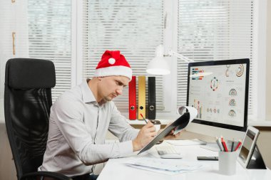 Pencere arka plan üzerinde ışık Office belgeleri ile modern monitör bilgisayarda çalışan yakışıklı ciddi ve dalmış iş adamı masa başında oturan gömlek ve kırmızı Noel şapkalı. Yeni yıl.