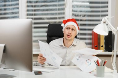 Yeni yıl bilgisayarda çalışan modern monitör, lamba, belge penceresi arka plan üzerinde ışık Office ile yakışıklı şok ve çapraşık iş adamı masa başında oturan gömlek, kırmızı Noel şapkalı.
