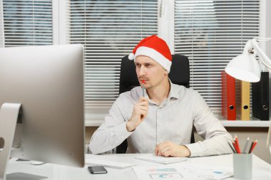Yeni yıl bilgisayarda çalışan modern monitör, lamba, belge penceresi arka plan üzerinde ışık Office ile yakışıklı ciddi ve dalmış iş adamı masa başında oturan gömlek, kırmızı Noel şapkalı.