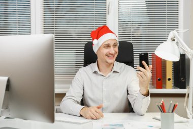 Eğlence gülümseyen yakışıklı selfie cep telefonu, bilgisayar, yeni yıl çalıştığı modern monitör, lamba, ışık Office belgeleri ile yapıyor iş adamı masa başında oturan gömlek, kırmızı Noel şapkalı.