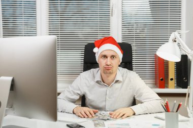 Yeni yıl bilgisayarda çalışan modern monitör, lamba, ışık Office belgeleri ile üzgün, şok ve çapraşık iş adamı ile çok nakit para, Masa başında oturan gömlek, kırmızı Noel şapkalı.